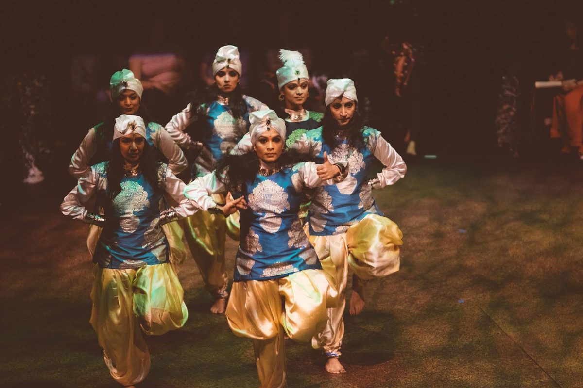 Six dancers in blue-and-silver tunics, gold trousers and white turbans hold a tightly grouped formation on a dimly lit stage; one kneels at the center while the others frame her.